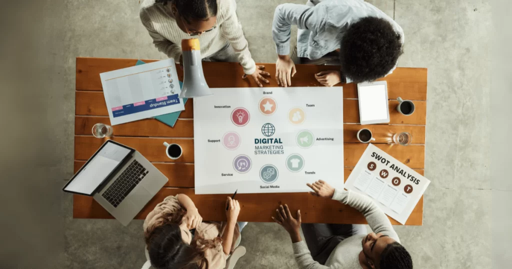 Profissionais reunidos ao redor de uma mesa desenvolvendo um planejamento de marketing com análise SWOT, gráficos e estratégias digitais.
